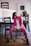 A woman wearing a pink suit and white blouse is seated on a chair in a casual pose. She has dark hair pulled back in a ponytail and is wearing black ankle boots. The setting appears to be a home or studio with wooden furniture, including chairs and a table. There are framed pictures on the wall and a plant in a woven basket on the table.