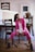 A woman wearing a pink suit and white blouse is seated on a chair in a casual pose. She has dark hair pulled back in a ponytail and is wearing black ankle boots. The setting appears to be a home or studio with wooden furniture, including chairs and a table. There are framed pictures on the wall and a plant in a woven basket on the table.