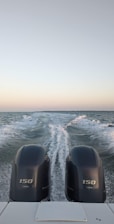 Two boat engines in the foreground create a trail of white foam on a vast, calm body of water under a clear sky. The horizon is visible in the distance, showing a subtle gradient from pale blue to soft pink.