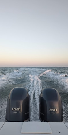 Two boat engines in the foreground create a trail of white foam on a vast, calm body of water under a clear sky. The horizon is visible in the distance, showing a subtle gradient from pale blue to soft pink.