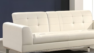 A modern, white, upholstered sofa with a minimalist design and tufted backrest sits in a room with a neutral color scheme. The background features a dark blue wall and contrasting white elements, creating a clean and contemporary aesthetic.