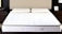 A neatly made bed features a white mattress with two cream-colored pillows. The mattress has a subtle pattern and appears supportive. Situated in a modern bedroom, the bed is flanked by dark wooden nightstands. An elegant, minimalist decor style is evident in the room.