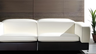 A modern white sofa with clean lines sits against a textured wall. A potted plant is situated to the right, adding a touch of greenery to the minimalistic setting.