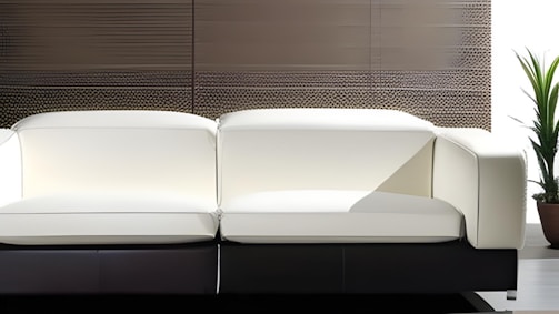 A modern white sofa with clean lines sits against a textured wall. A potted plant is situated to the right, adding a touch of greenery to the minimalistic setting.