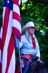 A person wearing a cowboy hat and western attire holds a large American flag. The individual has long hair and is dressed in a light blue shirt with a red scarf. A large decorative belt buckle is visible, and they appear to be sitting on a horse. The background is filled with green foliage.