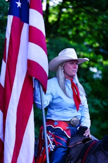 A person wearing a cowboy hat and western attire holds a large American flag. The individual has long hair and is dressed in a light blue shirt with a red scarf. A large decorative belt buckle is visible, and they appear to be sitting on a horse. The background is filled with green foliage.