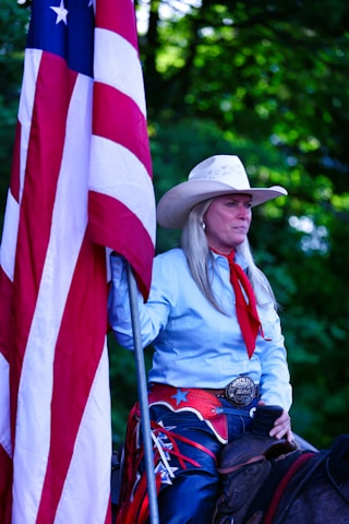 A person wearing a cowboy hat and western attire holds a large American flag. The individual has long hair and is dressed in a light blue shirt with a red scarf. A large decorative belt buckle is visible, and they appear to be sitting on a horse. The background is filled with green foliage.