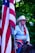 A person wearing a cowboy hat and western attire holds a large American flag. The individual has long hair and is dressed in a light blue shirt with a red scarf. A large decorative belt buckle is visible, and they appear to be sitting on a horse. The background is filled with green foliage.