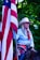 A person wearing a cowboy hat and western attire holds a large American flag. The individual has long hair and is dressed in a light blue shirt with a red scarf. A large decorative belt buckle is visible, and they appear to be sitting on a horse. The background is filled with green foliage.