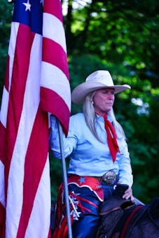 A person wearing a cowboy hat and western attire holds a large American flag. The individual has long hair and is dressed in a light blue shirt with a red scarf. A large decorative belt buckle is visible, and they appear to be sitting on a horse. The background is filled with green foliage.