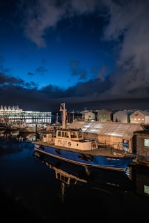 A pilot boat is docked at a marina under a dark, cloudy sky. The boat is predominantly blue and white, with the word 'PILOT' emblazoned on its side. Nearby, several other boats and buildings are visible, illuminated by soft lighting. The reflection of the boat can be seen in the calm water, adding to the serene ambiance. In the background, there are bright lights from a nearby structure or possibly a larger ship, contrasting with the deep blue of the sky.