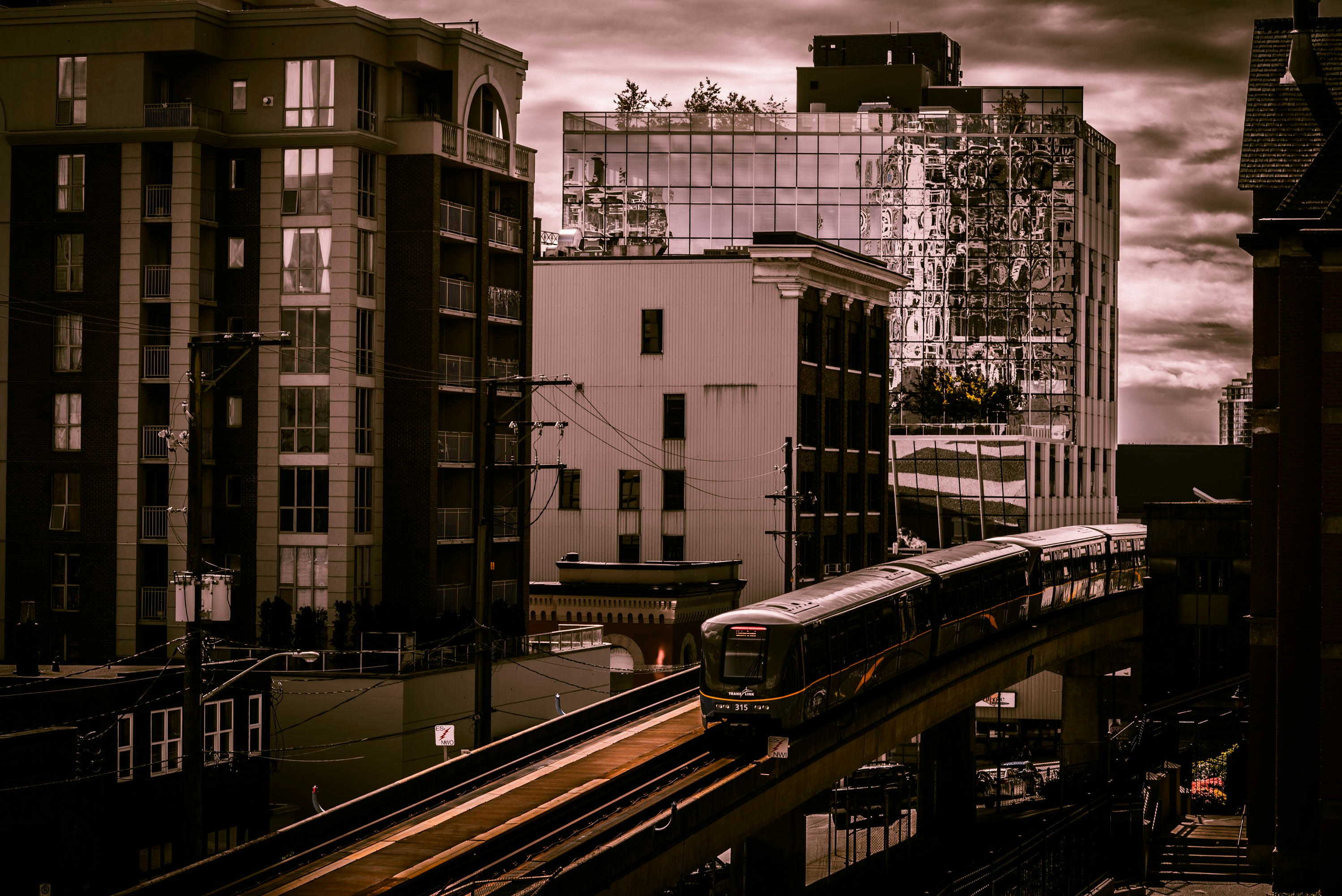 A modern cityscape featuring several tall buildings with reflective glass windows under a dramatic sky. An elevated train is traveling along a track in the foreground, weaving through the urban environment. The lighting creates a moody atmosphere, with an emphasis on the architectural details of the buildings.