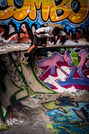A person in casual attire is performing a skateboard trick on the edge of a graffiti-covered skate park. The walls are adorned with vibrant and colorful street art featuring bold patterns and letters.