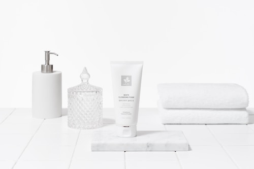 A clean and minimalist arrangement featuring a white tube of cleansing foam on a marble slab, accompanied by a white pump dispenser, a crystal-like jar with a lid, and a stack of white towels. The setting gives an impression of a bathroom or spa with a focus on skincare products.