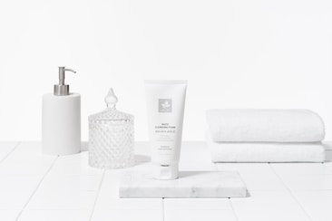A clean and minimalist arrangement featuring a white tube of cleansing foam on a marble slab, accompanied by a white pump dispenser, a crystal-like jar with a lid, and a stack of white towels. The setting gives an impression of a bathroom or spa with a focus on skincare products.