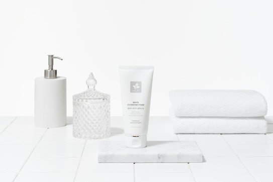 A clean and minimalist arrangement featuring a white tube of cleansing foam on a marble slab, accompanied by a white pump dispenser, a crystal-like jar with a lid, and a stack of white towels. The setting gives an impression of a bathroom or spa with a focus on skincare products.