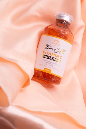A bottle labeled 'Stem Cell Vitamin C Whitening' with the words 'Pure Fresh Botox Ampoule' is placed on soft, peach-colored fabric. The fabric has gentle folds creating a subtle, luxurious texture. The bottle is clear with an orange liquid inside, and it has a white and orange label with elegant typography.