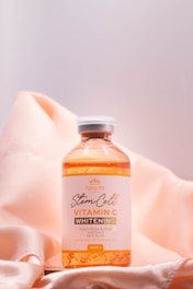 A bottle of vitamin C whitening product is positioned on a peach-colored, silky fabric backdrop. The label on the bottle indicates it contains stem cell ingredients for skin rejuvenation.