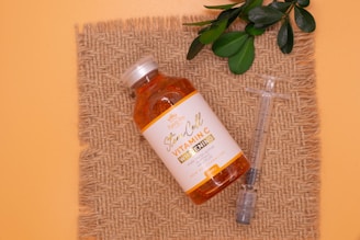 A bottle labeled 'Stem Cell Vitamin C Whitening' is placed on a piece of burlap fabric. Beside it lies a syringe, and some green leaves are placed above the bottle, creating a natural and organic feel. The background is a soft peach color.