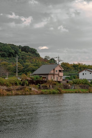 A house with a green roof is situated near a body of water, surrounded by lush greenery. The sky is overcast with gray clouds, and the landscape includes hills covered in dense foliage.