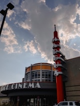 A cinema building with a prominent red and silver vertical sign is set against a partly cloudy sky. The building displays a rounded facade with large glass windows and the sign spells out letters vertically.
