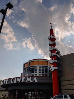 A cinema building with a prominent red and silver vertical sign is set against a partly cloudy sky. The building displays a rounded facade with large glass windows and the sign spells out letters vertically.