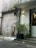 An exterior wall of a building features a mix of materials and textures, including faded paint and graffiti. Vegetation is visible in crevices and a planter near the steps. A light fixture illuminates the wall, and a door leads into the building.