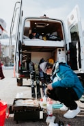A person is crouching next to a portable grill cooking food outside. Behind them is an open van with its back doors wide open, revealing an interior with a bed and equipment. Ski gear is leaning against the van, and another person is visible inside.