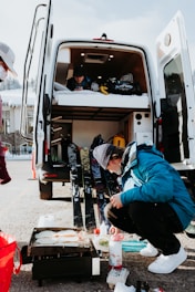 A person is crouching next to a portable grill cooking food outside. Behind them is an open van with its back doors wide open, revealing an interior with a bed and equipment. Ski gear is leaning against the van, and another person is visible inside.