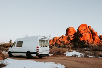 A white campervan is parked on a dirt road in a scenic area with large, reddish-orange rock formations in the background. Sparse snow patches are visible on the ground, and the overall landscape has a rugged, natural appearance. A few small trees and bushes are scattered around the rocks, under a clear sky at sunset or sunrise, casting warm light on the rocks.