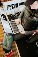 A person wearing casual outdoor clothing, including a padded jacket and cap, is sitting with a laptop on their lap. They are interacting with the laptop, possibly working or browsing. Nearby, various electronic devices and cables are visible, and the person’s backpack is placed beside them, indicating a mobile or on-the-go workspace.
