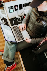 A person wearing casual outdoor clothing, including a padded jacket and cap, is sitting with a laptop on their lap. They are interacting with the laptop, possibly working or browsing. Nearby, various electronic devices and cables are visible, and the person’s backpack is placed beside them, indicating a mobile or on-the-go workspace.