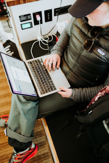 A person wearing casual outdoor clothing, including a padded jacket and cap, is sitting with a laptop on their lap. They are interacting with the laptop, possibly working or browsing. Nearby, various electronic devices and cables are visible, and the person&rsquo;s backpack is placed beside them, indicating a mobile or on-the-go workspace.