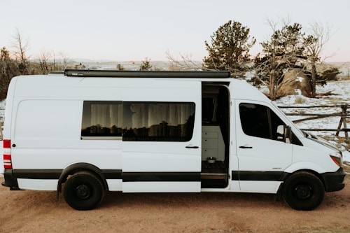 A white camper van with its side door open, parked on a dirt path. The interior can be seen slightly through the open door, revealing some cabinets. Snow covers the ground and trees in the background, with a wooden fence also visible. The sky is overcast with a soft light suggesting late afternoon or early evening.