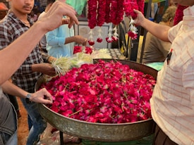 A bustling flower market scene with several vendors and buyers. A large metal container is filled with vibrant red flowers, possibly roses or hibiscus. People are holding and selecting flowers, with marigolds visible in one person's hands. Bundles of white flowers are also present. Hands are reaching in, likely preparing or purchasing flower garlands. Bright and colorful atmosphere.