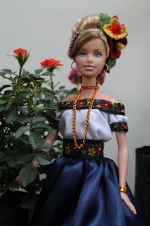 A doll dressed in traditional attire stands next to a small plant with blooming red roses. The doll is adorned in a white off-shoulder top with black and floral embroidered trim, a blue skirt, and gold jewelry, including a necklace, earrings, and bracelet. The hair is styled in an updo, decorated with vibrant flowers.