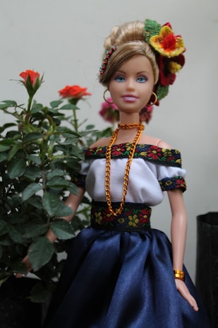 A doll dressed in traditional attire stands next to a small plant with blooming red roses. The doll is adorned in a white off-shoulder top with black and floral embroidered trim, a blue skirt, and gold jewelry, including a necklace, earrings, and bracelet. The hair is styled in an updo, decorated with vibrant flowers.