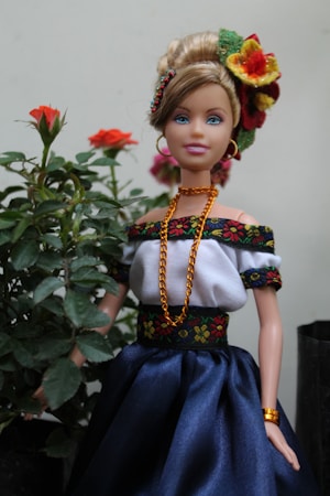A doll dressed in traditional attire stands next to a small plant with blooming red roses. The doll is adorned in a white off-shoulder top with black and floral embroidered trim, a blue skirt, and gold jewelry, including a necklace, earrings, and bracelet. The hair is styled in an updo, decorated with vibrant flowers.