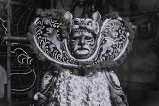 A person is wearing an elaborate costume featuring intricate patterns and designs resembling a lion, with a headdress that includes a mask covering the face. The costume is adorned with detailed artwork and floral elements.