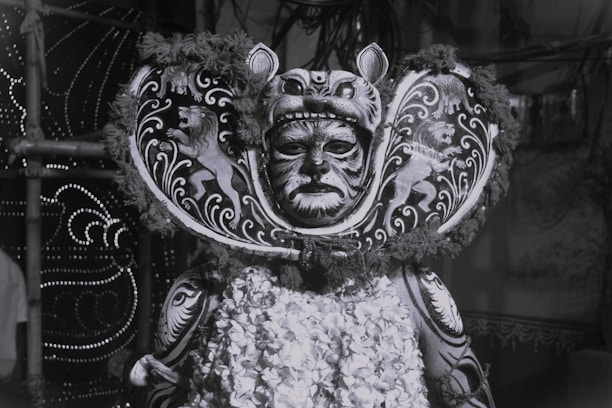 A person is wearing an elaborate costume featuring intricate patterns and designs resembling a lion, with a headdress that includes a mask covering the face. The costume is adorned with detailed artwork and floral elements.