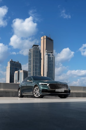 A sleek, modern car is parked on a rooftop with a backdrop of tall skyscrapers under a bright blue sky. The clouds are fluffy and create a vibrant atmosphere. The car's design is elegant and shiny, reflecting the sunlight.