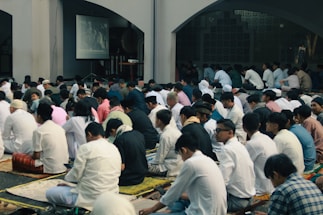 A large group of people, mostly men, are seated on the floor in a mosque-like setting. They are predominantly wearing white clothing and traditional caps. A screen displays an image in the background, suggesting an ongoing event or presentation. The atmosphere appears to be solemn and focused.