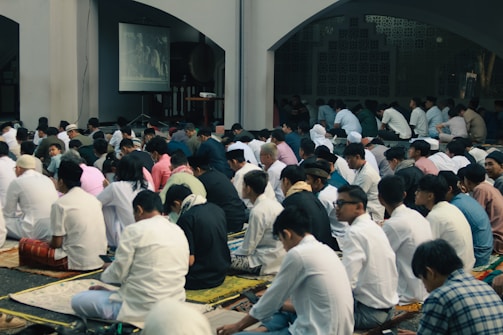 A large group of people, mostly men, are seated on the floor in a mosque-like setting. They are predominantly wearing white clothing and traditional caps. A screen displays an image in the background, suggesting an ongoing event or presentation. The atmosphere appears to be solemn and focused.
