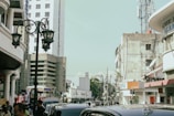 An urban street scene with several mid-rise buildings and a telecommunications tower in the background. The street is bustling with people and vehicles, and ornate street lamps are visible. One of the buildings has a sign with the letters 'BNI' and another one labeled 'BRAGA'.