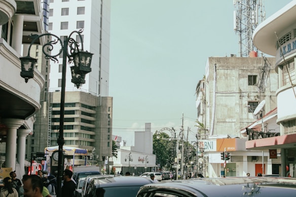 An urban street scene with several mid-rise buildings and a telecommunications tower in the background. The street is bustling with people and vehicles, and ornate street lamps are visible. One of the buildings has a sign with the letters 'BNI' and another one labeled 'BRAGA'.