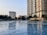 A large outdoor swimming pool surrounded by tall modern buildings and palm trees. Several people are enjoying the water, with some lounging or swimming. The sky is clear, suggesting a warm and pleasant day.