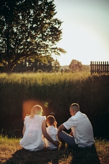 A family of three, composed of two adults and a child, sits on the grass near a small body of water, during what appears to be sunset. The sunlight creates a warm glow, with a large tree nearby casting shadows on the ground. The scene conveys a peaceful and serene atmosphere.