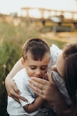 A woman gently embraces a young boy, who is looking downwards with a serene expression. They are surrounded by green grass, with a blurred background that suggests an outdoor setting. The woman's hand, featuring a simple ring, is tenderly touching the boy's face.