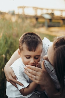 A woman gently embraces a young boy, who is looking downwards with a serene expression. They are surrounded by green grass, with a blurred background that suggests an outdoor setting. The woman's hand, featuring a simple ring, is tenderly touching the boy's face.
