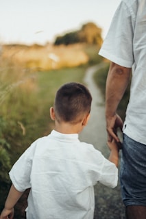 A child and an adult are walking hand in hand along a path surrounded by grass. They are dressed in casual summer clothing, and the image is taken from behind, focusing on their close bond.